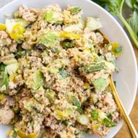 Salmon salad with pickles, avocado, fresh herbs and more mixed up in a white serving bowl with a golden spoon, ready to eat.