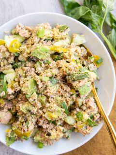 Salmon salad with pickles, avocado, fresh herbs and more mixed up in a white serving bowl with a golden spoon, ready to eat.