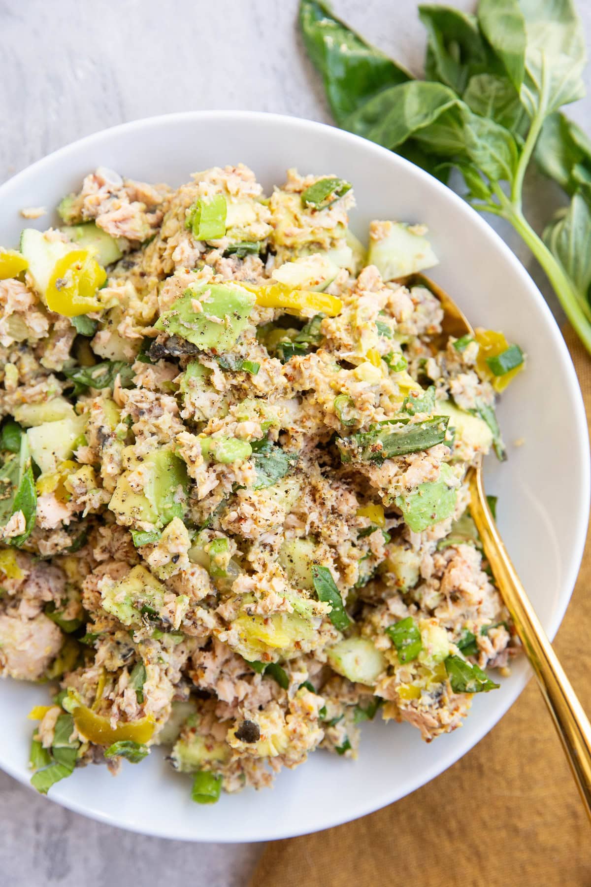 Salmon salad with pickles, avocado, fresh herbs and more mixed up in a white serving bowl with a golden spoon, ready to eat.