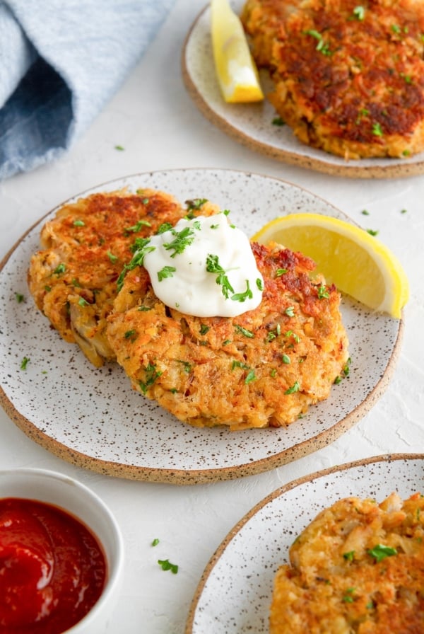 Three appetizer plates of crab cakes with tartar sauce, cocktail sauce, and lemon wedges.