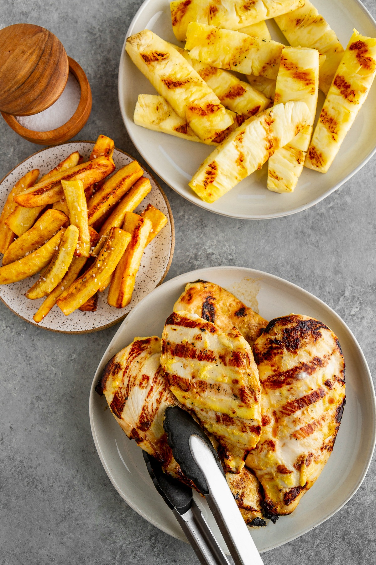 Grilled chicken and grilled pineapple on plates and a plate to the side of fried plantains.