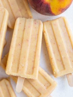 Pile of peach popsicles on a white background with a fresh peach to the side.