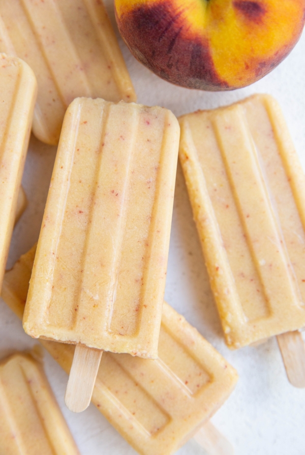 Pile of peach popsicles on a white background with a fresh peach to the side.