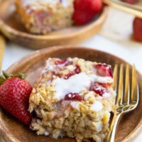 Two wooden plates of strawberry baked oatmeal with a glaze on top and golden forks with fresh strawberries to the side.