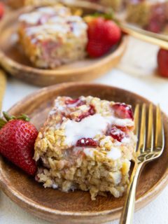 Two wooden plates of strawberry baked oatmeal with a glaze on top and golden forks with fresh strawberries to the side.