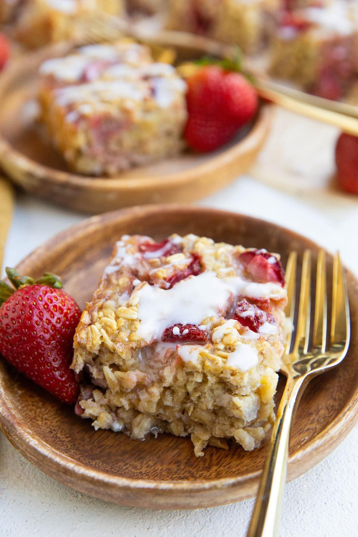 Two wooden plates of strawberry baked oatmeal with a glaze on top and golden forks with fresh strawberries to the side.