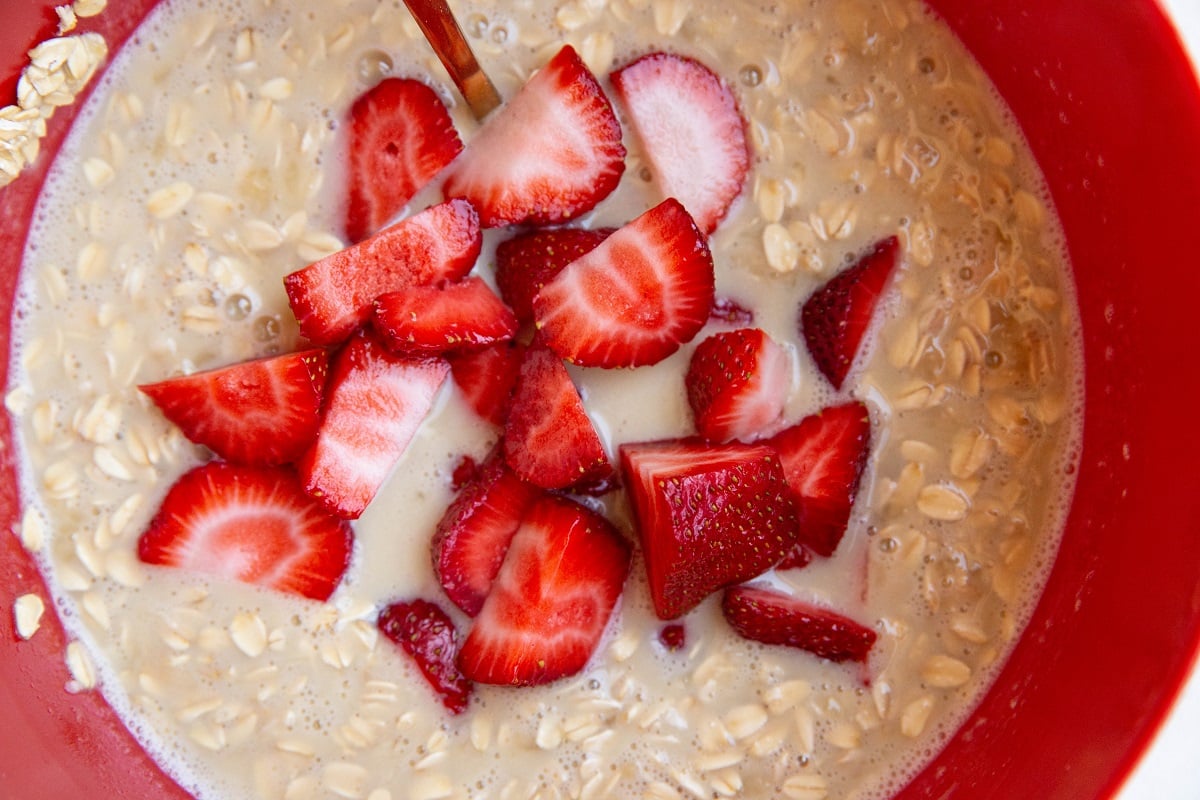 Oatmeal mixture in a red mixing bowl with strawberries on top, ready to be mixed in.