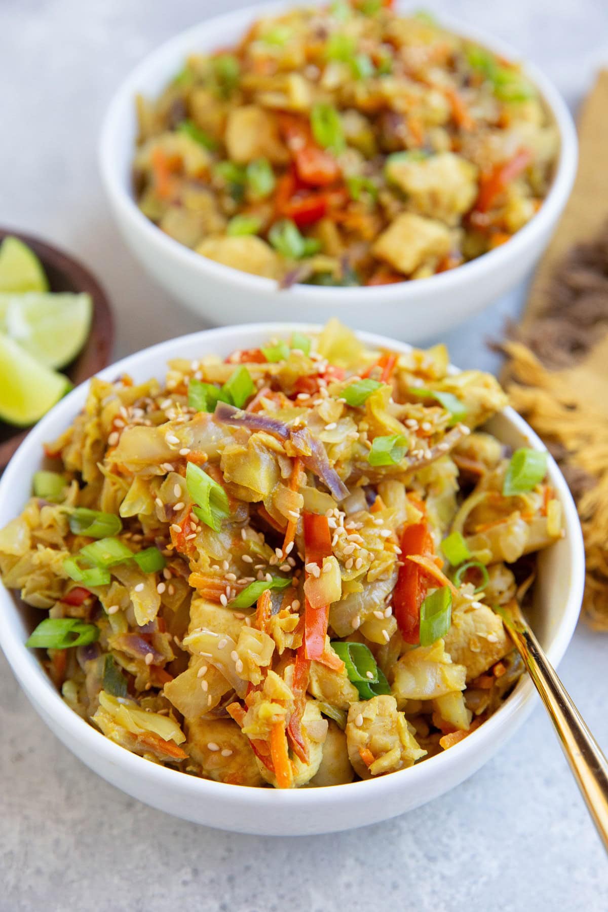 Two white bowls of Thai chicken with vegetables. A gold fork inside of the first bowl with a bowl of lime wedges in the background.