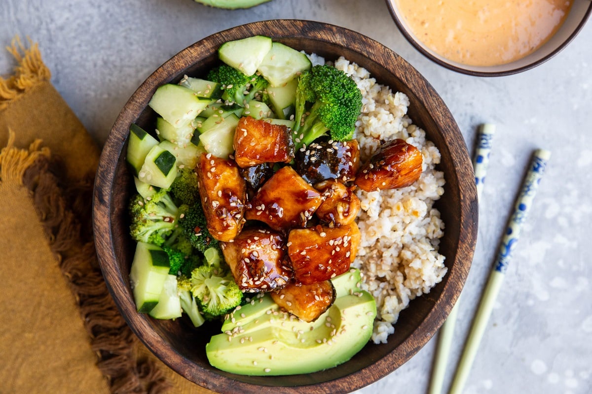 Wooden bowl of salmon bites with brown rice, broccoli, cucumber salad and avocado.