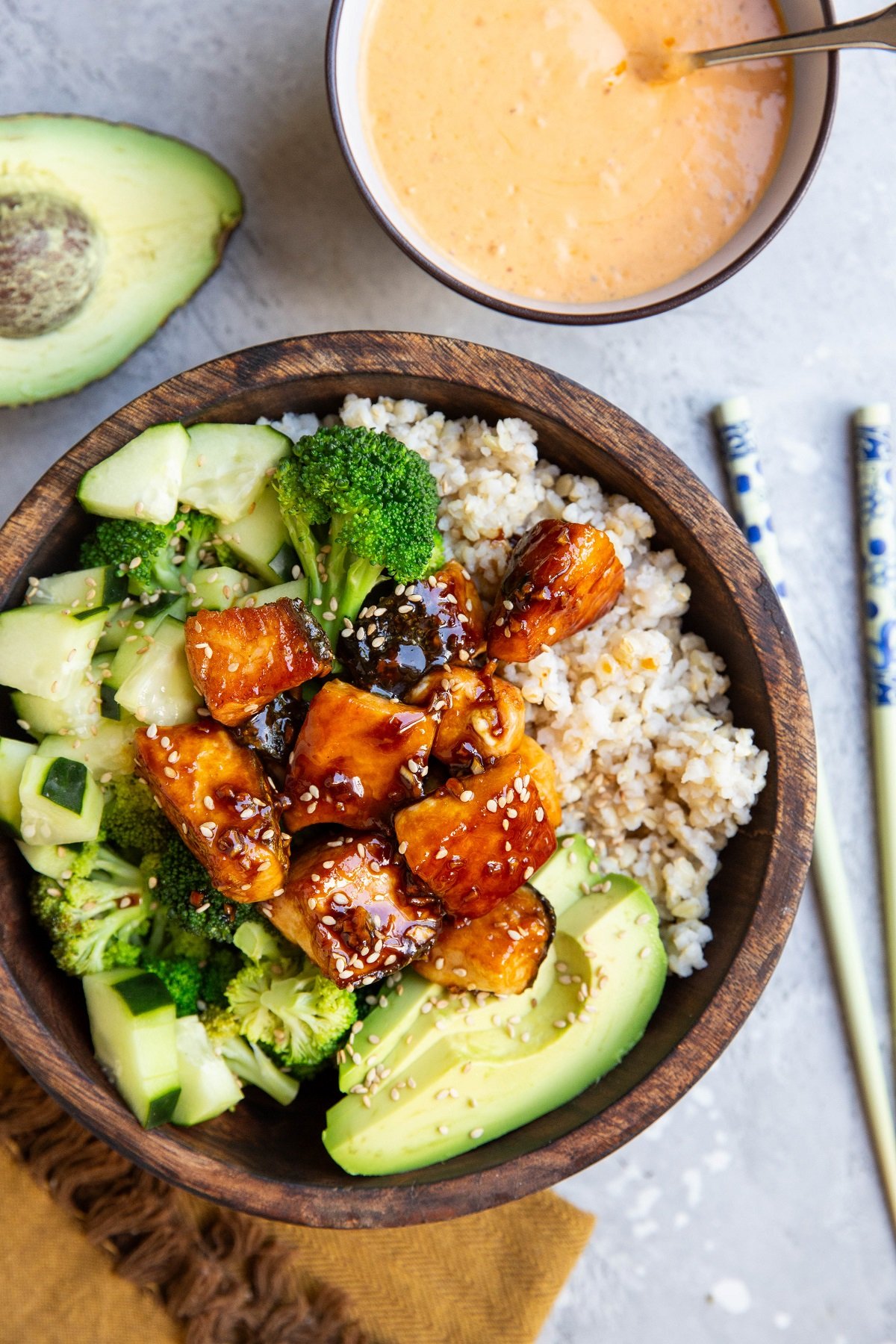 Bowl of salmon bites and vegetables with bang bang sauce to the side. Avocado, napkin and chop sticks to the side.