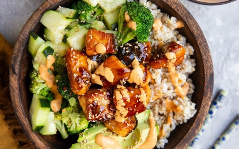 Wood bowl of brown rice, salmon bites, broccoli, and avocado and cucumbers.