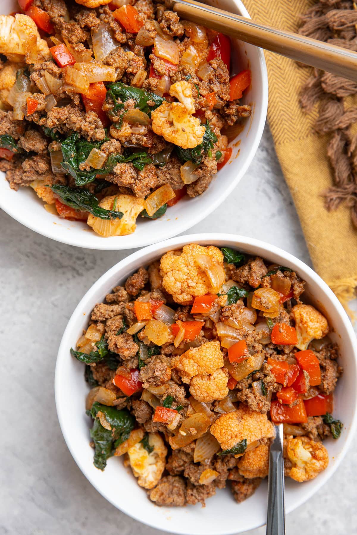 Two white bowls full of ground beef and vegetables with a golden napkin to the side.