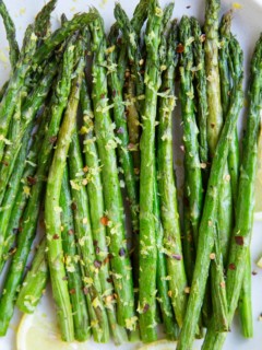 White plate of asparagus with lemon zest and pepper on top and fresh lemons to the side.