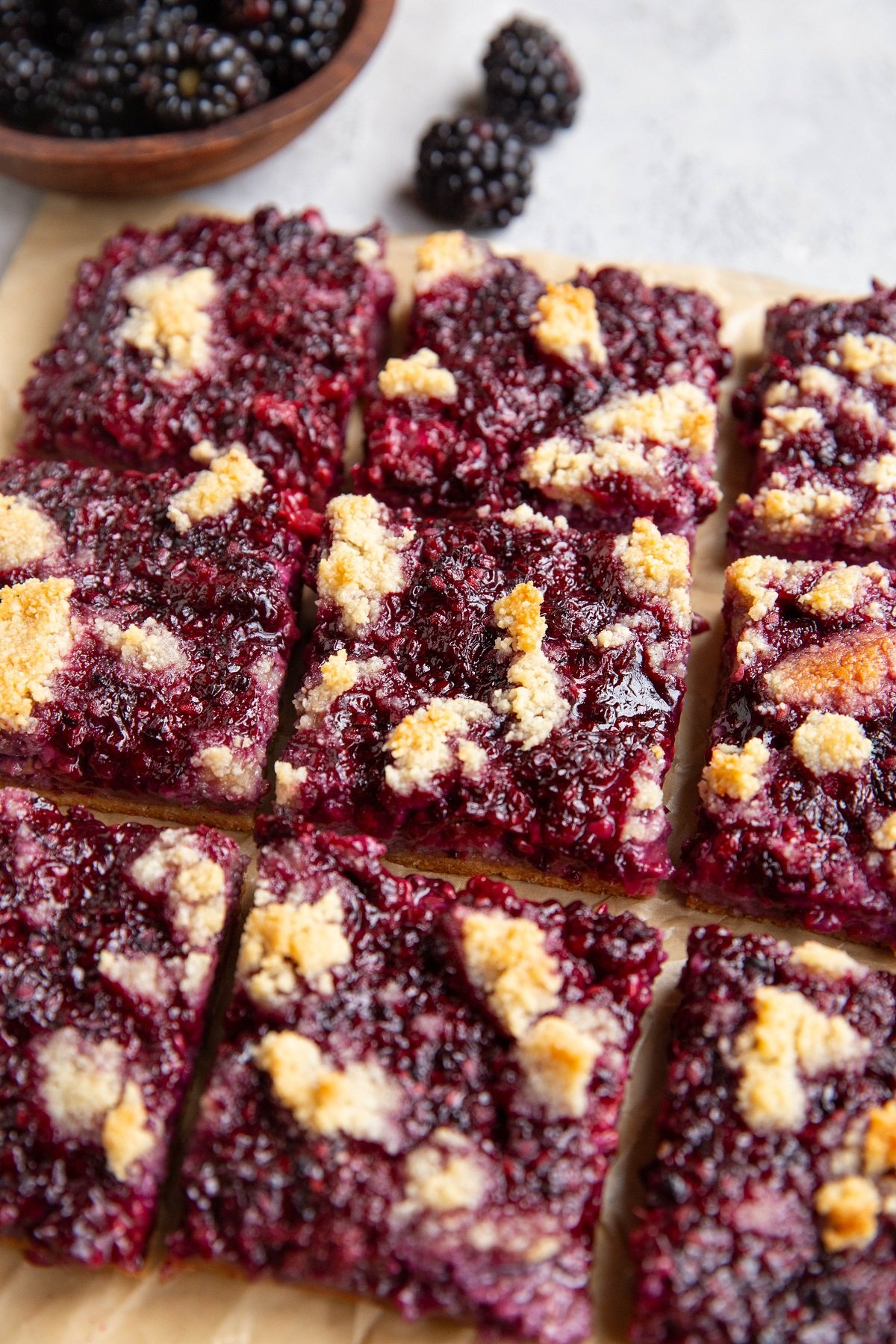 Blackberry bars on parchment paper with a bowl of blackberries in the background.