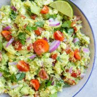 Bowl of chunky avocado dip with fresh tomatoes in a blue rimmed bowl with sliced limes to the side.