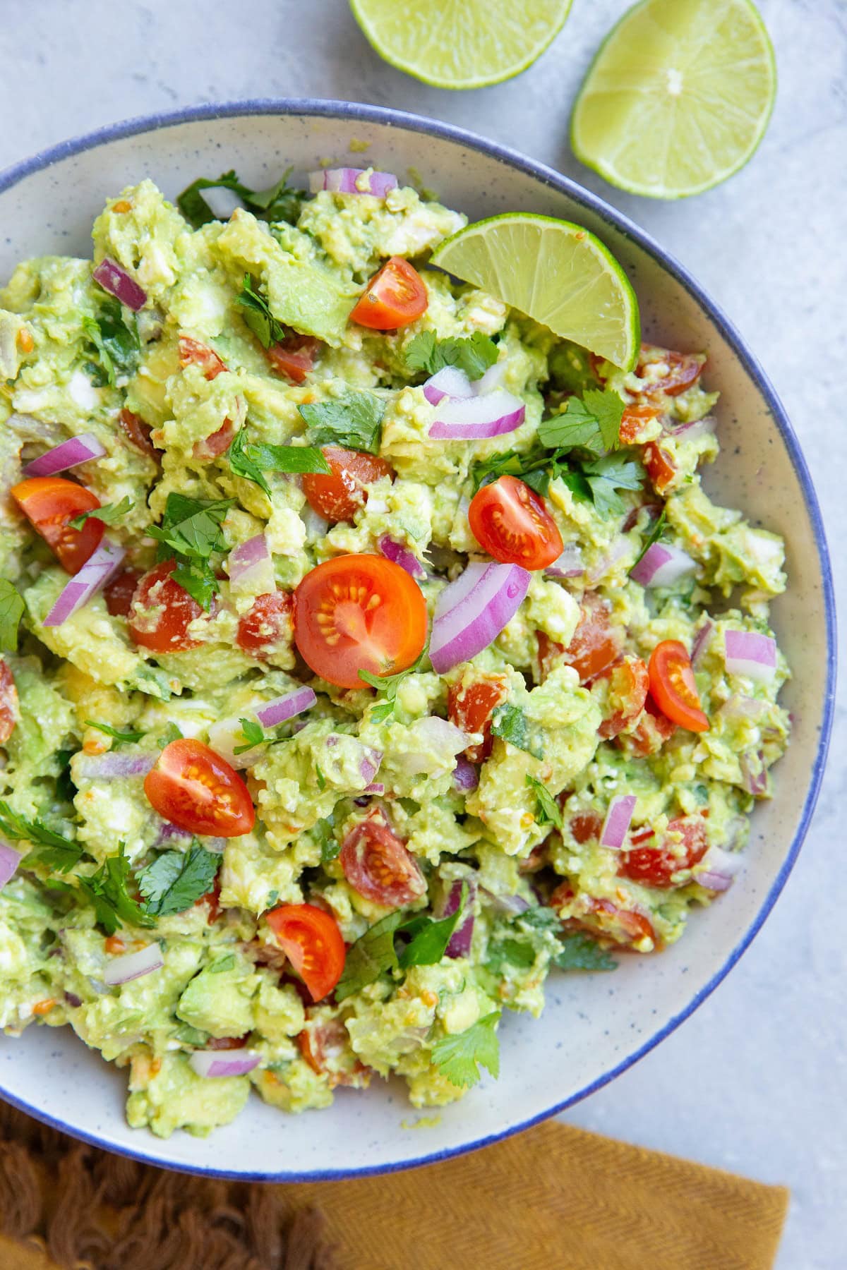 Bowl of chunky avocado dip with fresh tomatoes in a blue rimmed bowl with sliced limes to the side.