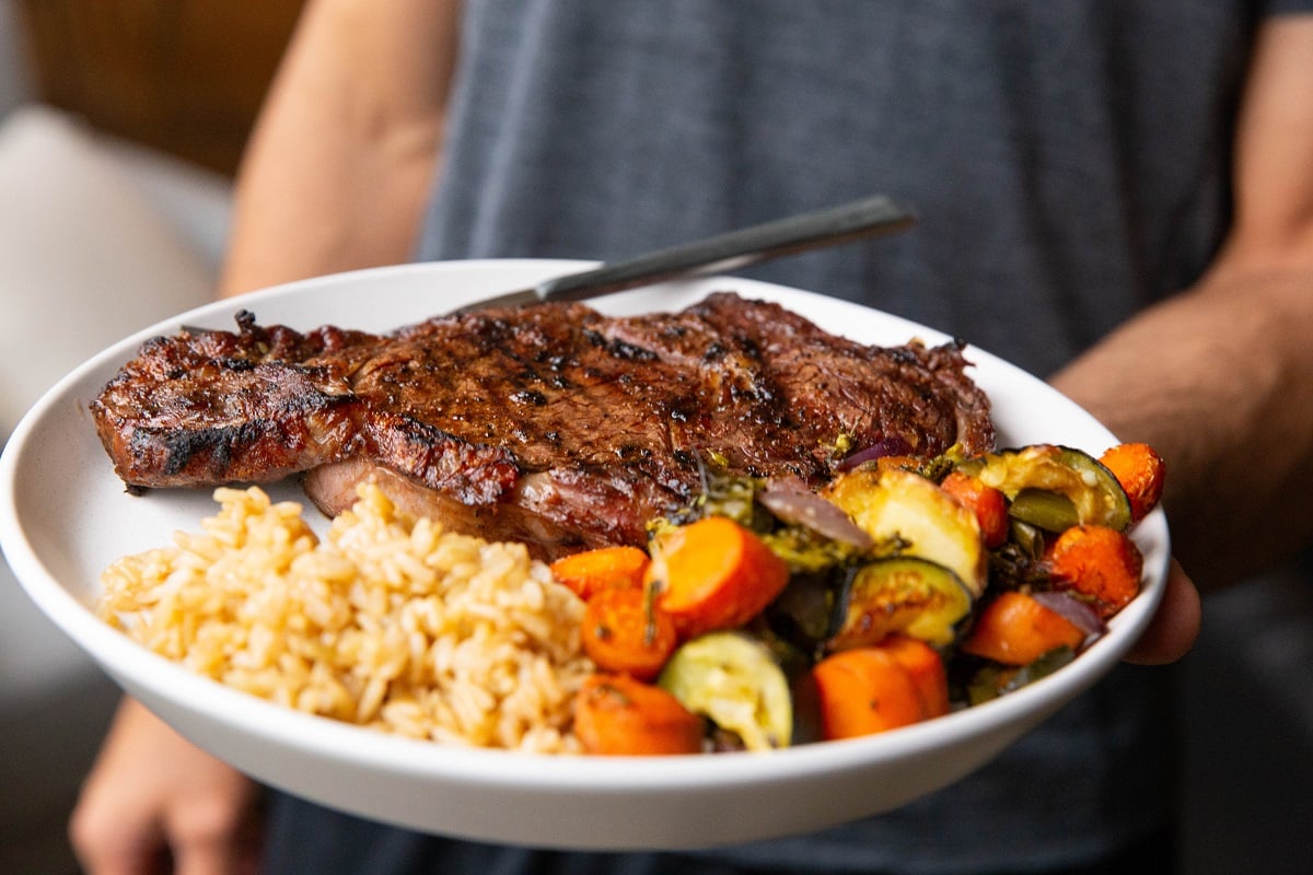 Man holding a plate with grilled ribeye, roasted vegetables and rice.