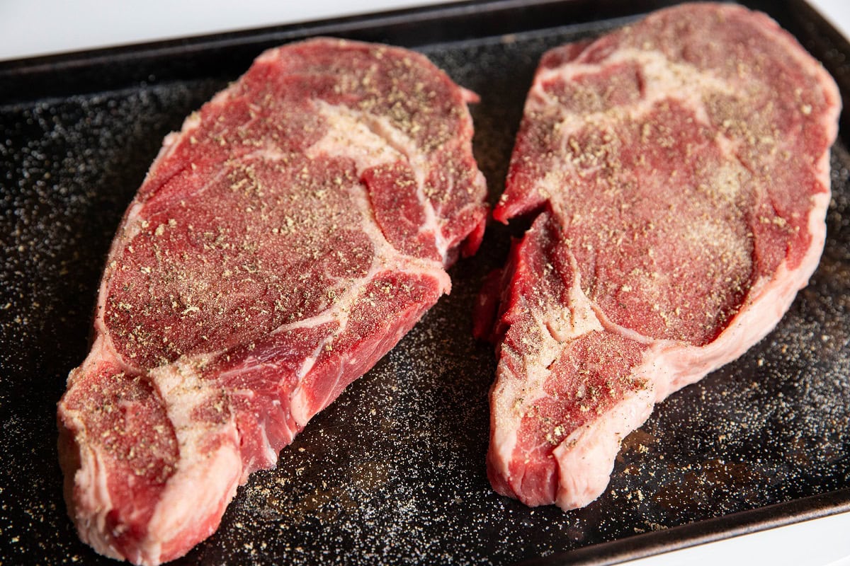 Two boneless ribeye steaks on a baking sheet, sprinkled with sea salt, garlic powder, and pepper.