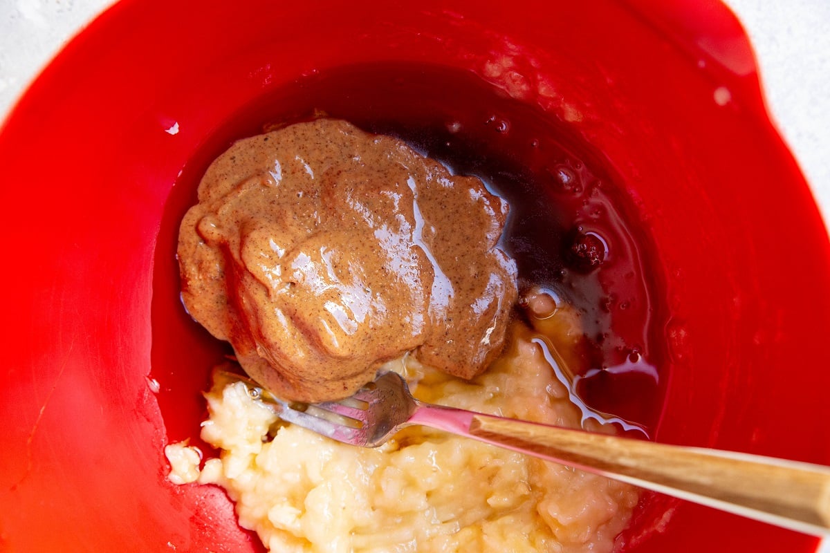 Mashed bananas, almond butter and pure maple syrup in a red mixing bowl, ready to be stirred together.