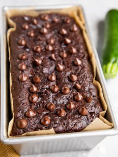 Chocolate zucchini bread in a loaf pan fresh out of the oven with a zucchini to the side.