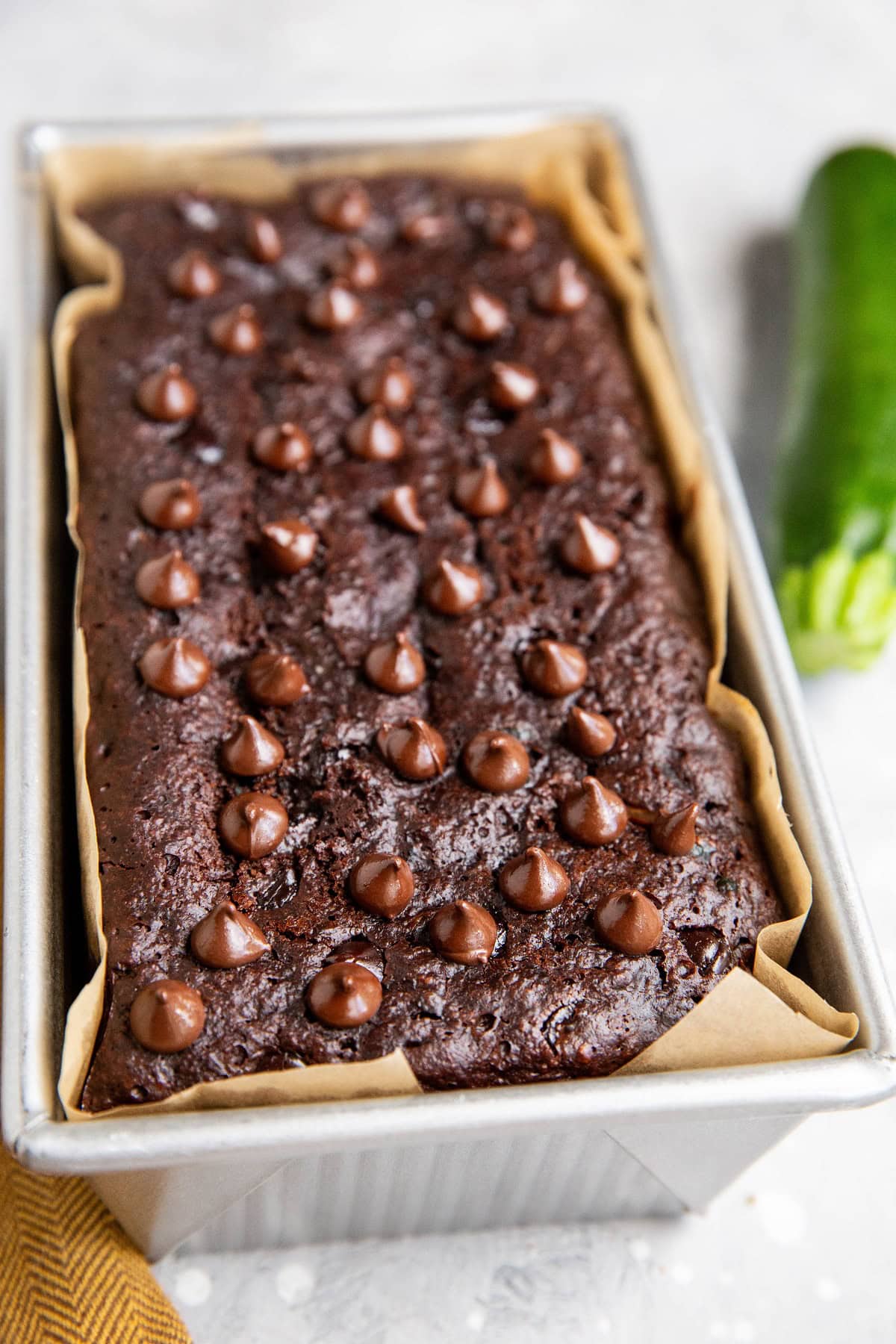 Chocolate zucchini bread in a loaf pan fresh out of the oven with a zucchini to the side.