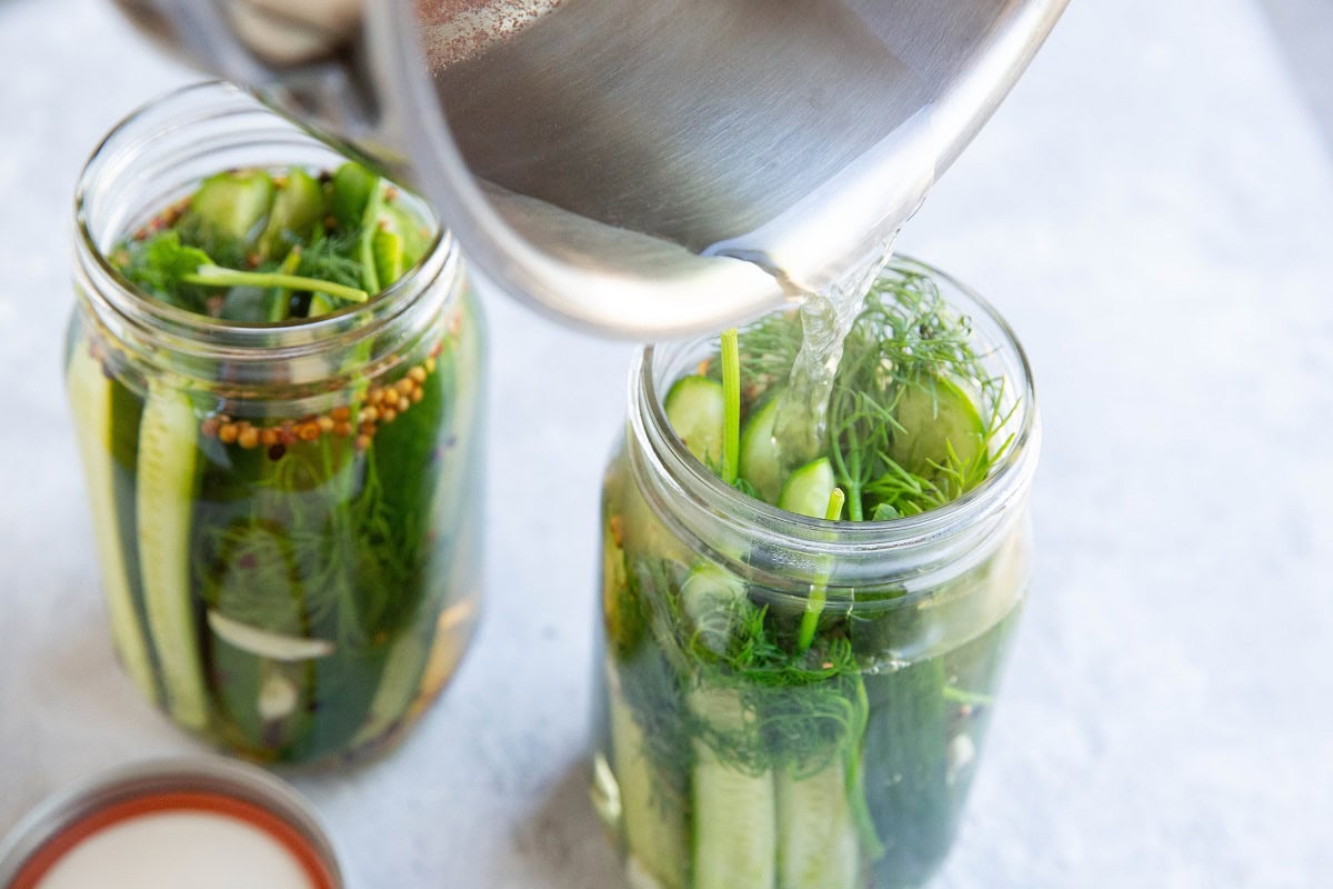 Pouring vinegar mixture into two jars full of pickle ingredients.