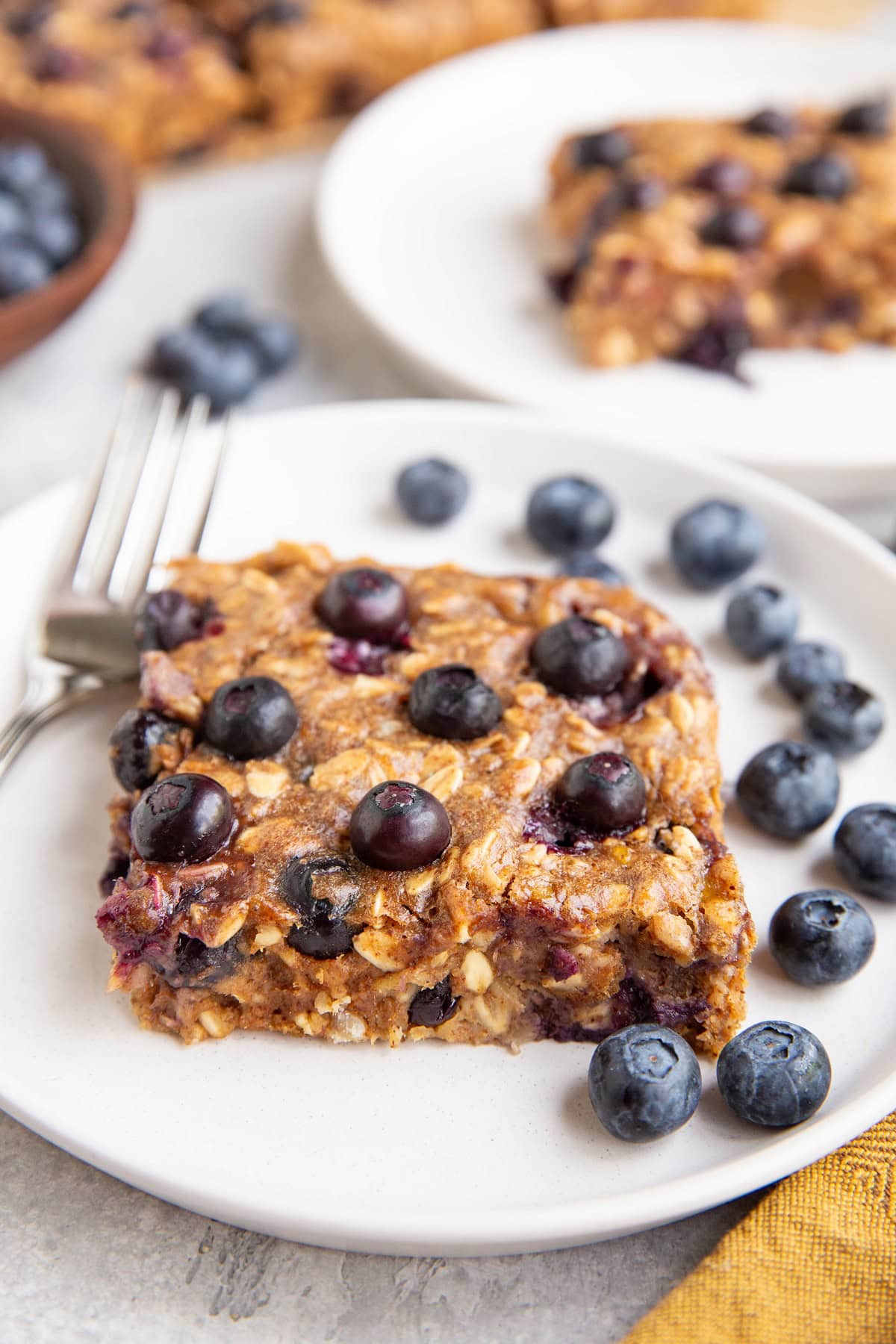 close up of a blueberry bar on a white plate with another plate of blueberry bar in the background.