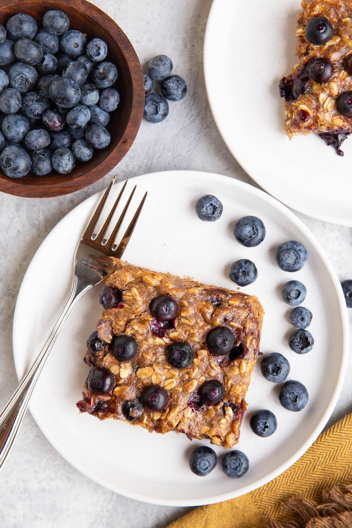 Two white plates with slices of blueberry bars and fresh blueberries all around.