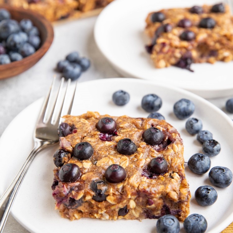 Two white plates of blueberry breakfast bars with fresh berries all around.