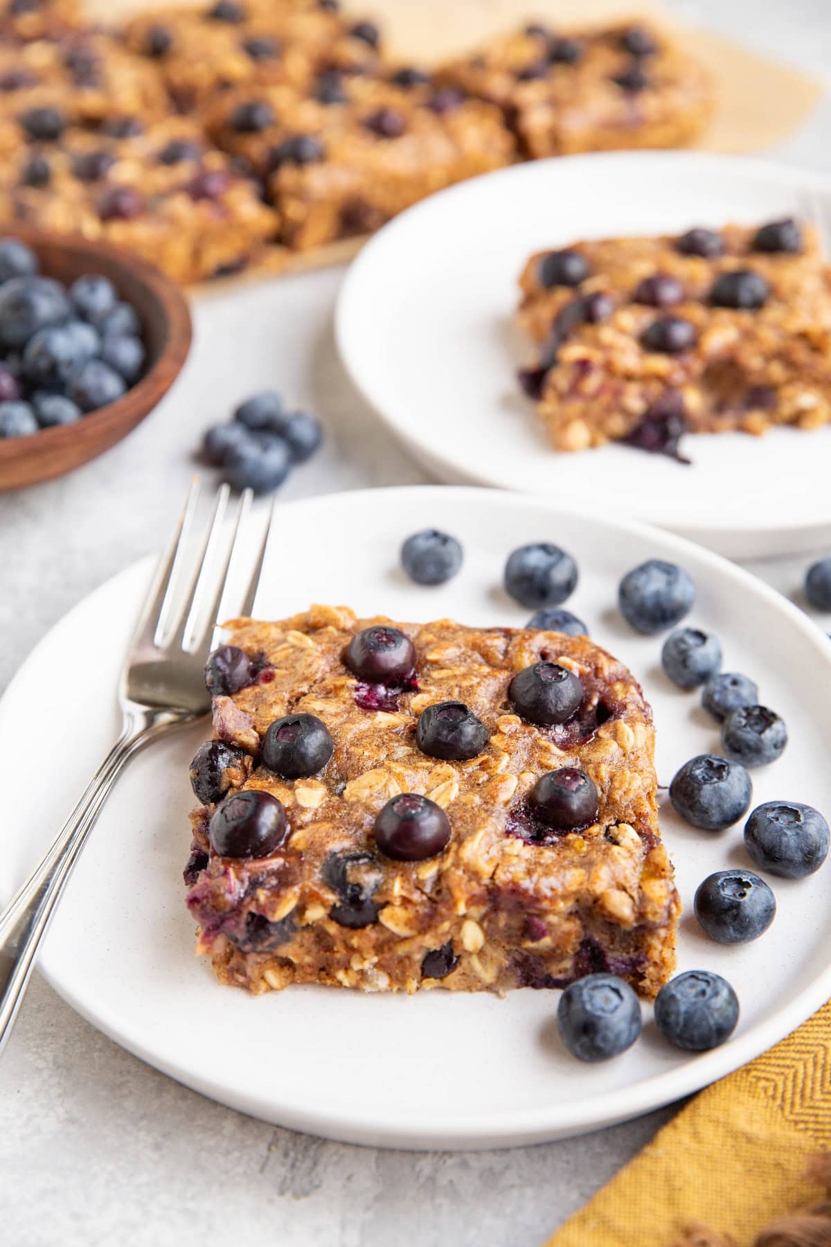 Two white plates of blueberry breakfast bars with fresh berries all around.