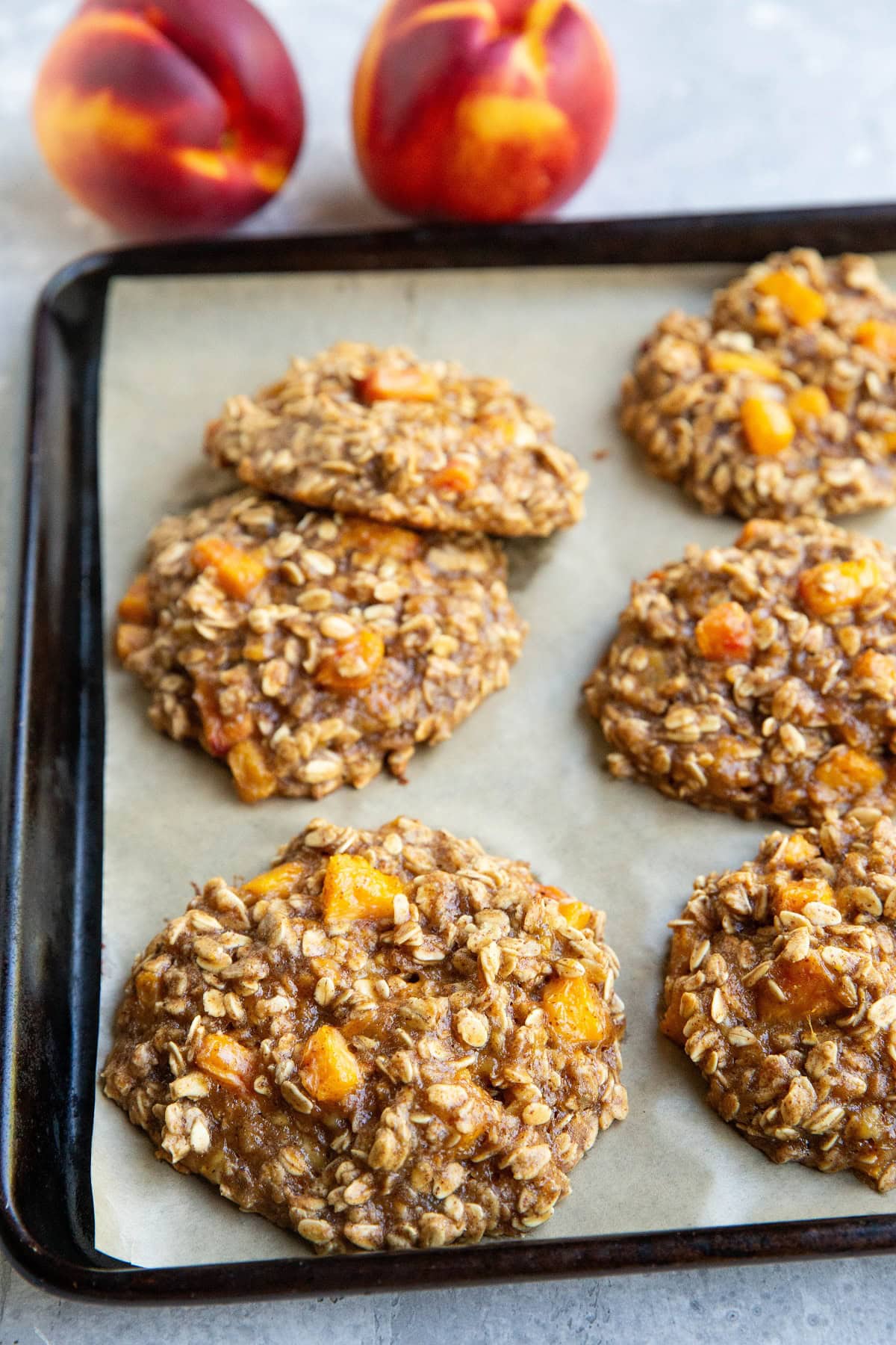 Baking sheet with large peach cookies on it, fresh out of the oven and two fresh peaches in the background.