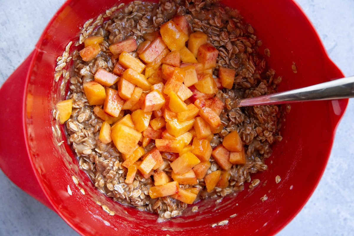 Red mixing bowl of an oatmeal mixture with peaches ready to be mixed in