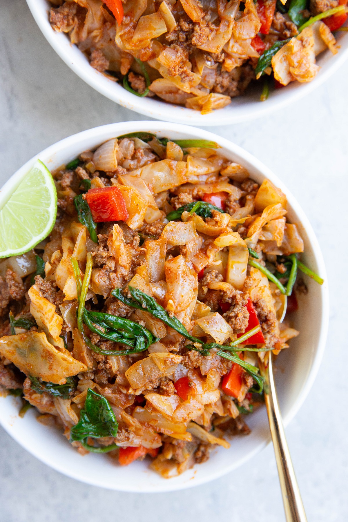 Two white bowls full of ground beef and cabbage with a slice of lime and a gold spoon.