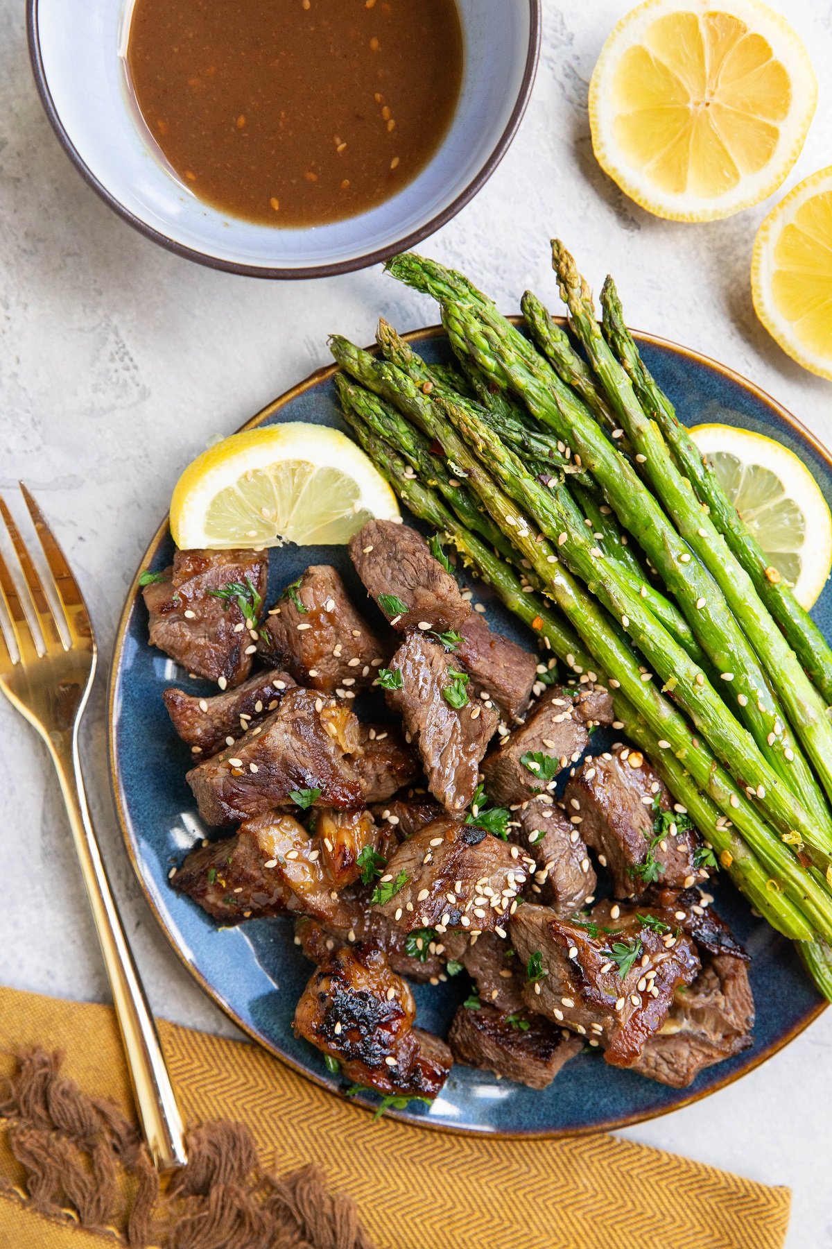 Blue plate of steak bites and asparagus, ready to serve. a bowl of teriyaki sauce to the side and fresh lemons. gold fork and napkin.