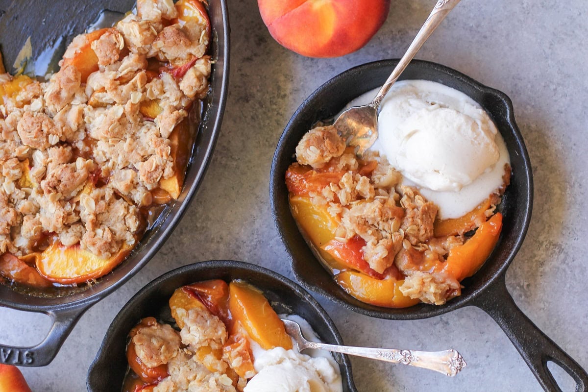 Two servings of peach cobbler in separate skillets with ice cream and the rest of the peach cobbler to the side.