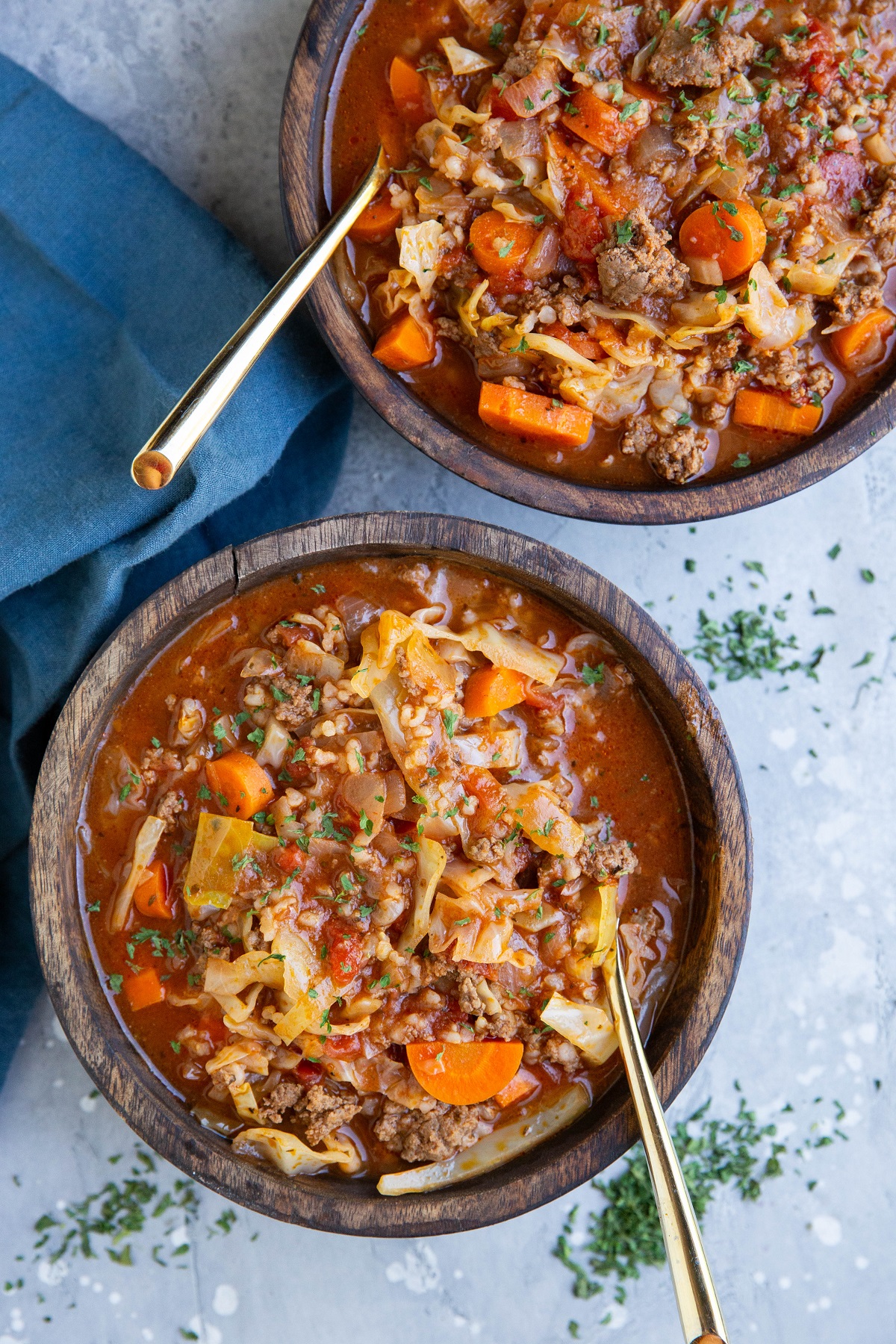 Two wooden bowls of cabbage roll soup with golden spoons and a blue napkin to the side.