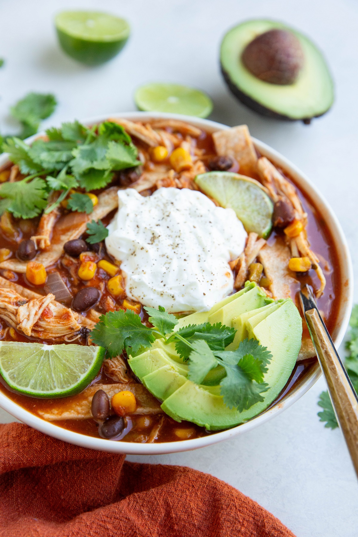 Big white bowl of chicken tortilla soup with toppings and avocado and limes in the background.