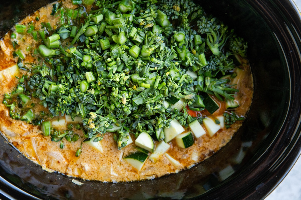 Fresh vegetables on top of red Thai curry in a crock pot, ready to be mixed in and cooked.