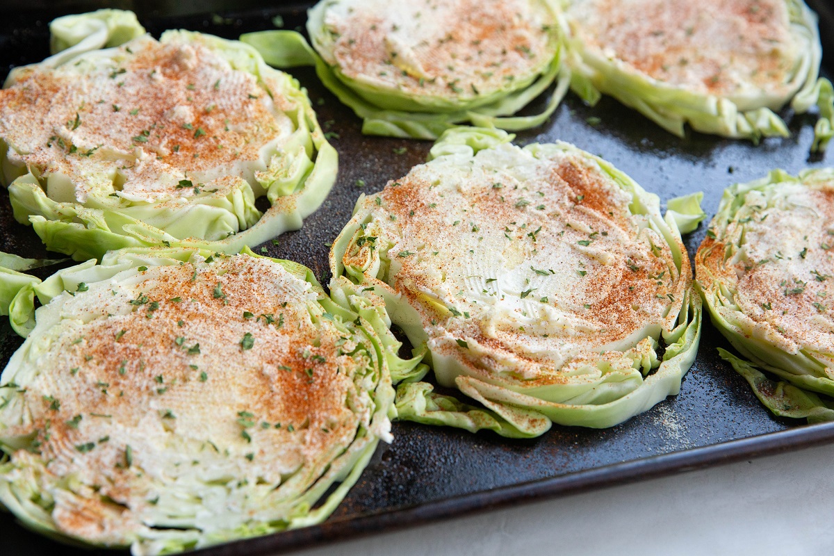 Raw rounds of cabbage on a baking sheet sprinkled with seasonings, ready to go into the oven.