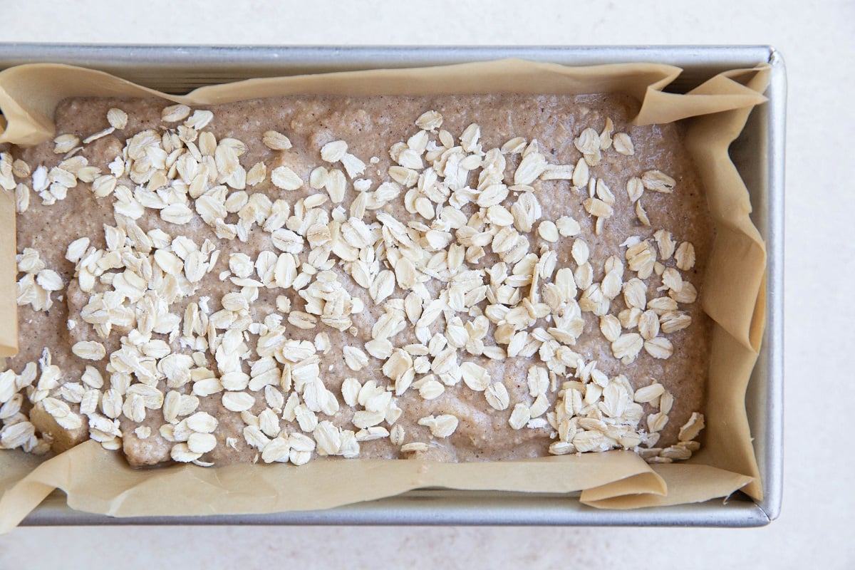 Loaf pan lined with parchment paper with apple oatmeal bread batter inside, ready to bake.