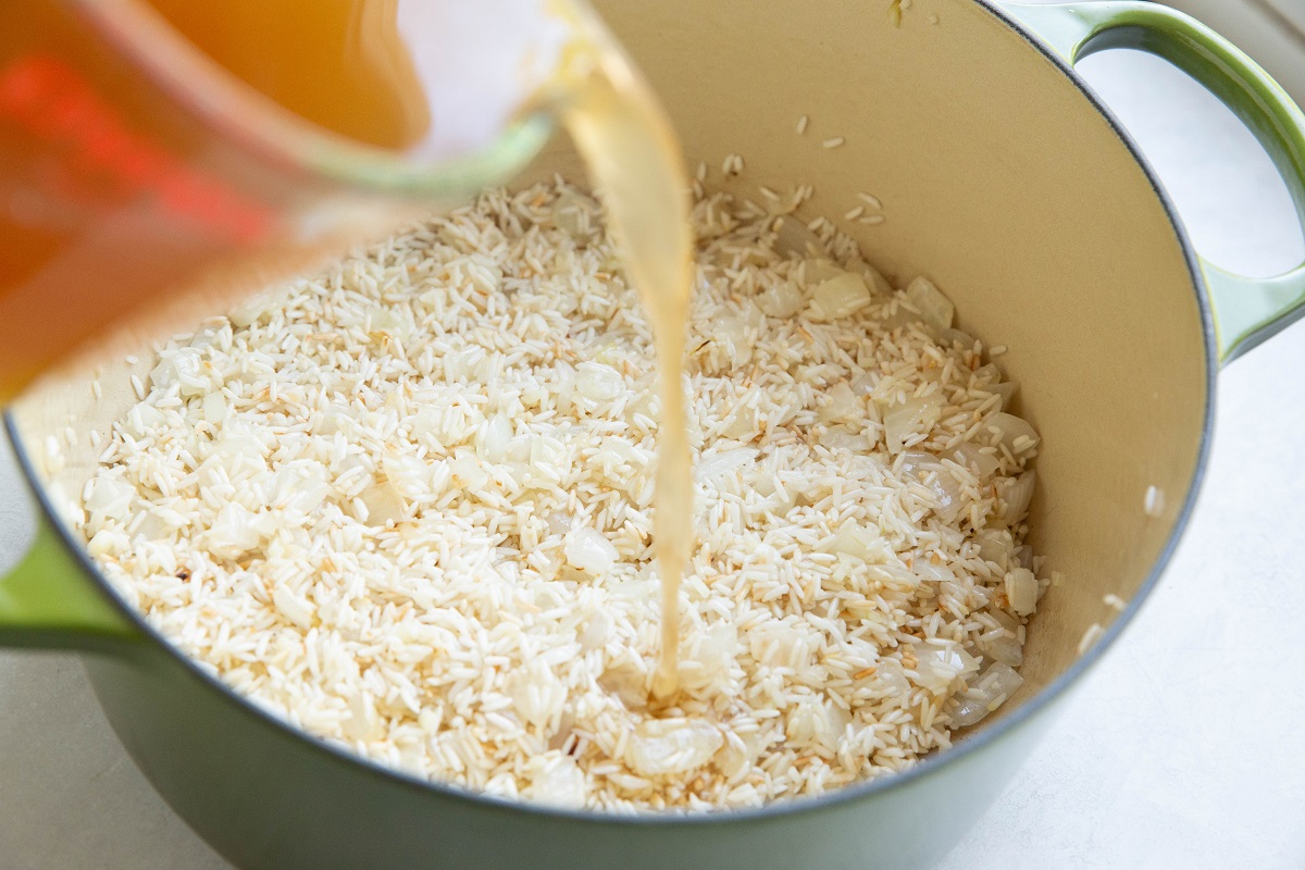 Pouring chicken broth into a large pot with rice, onion and garlic.