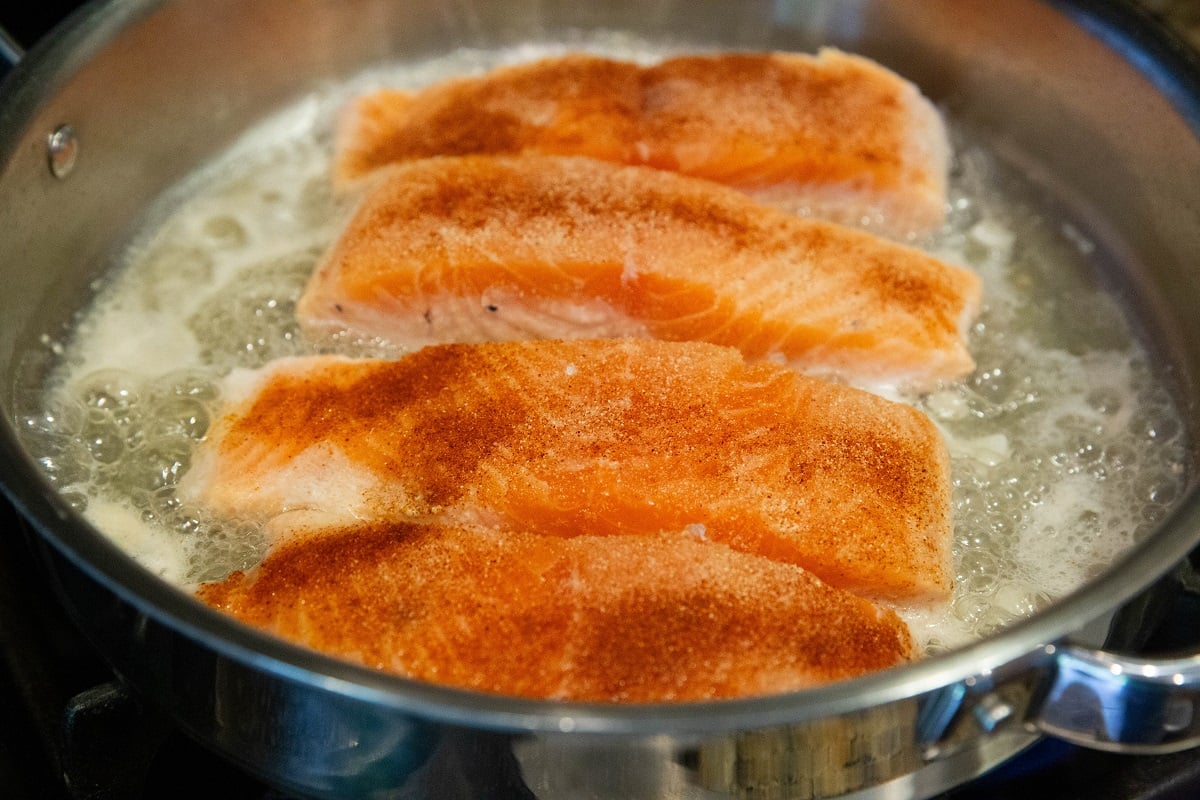Four salmon fillets cooking in poaching liquid in a skillet to make poached salmon.