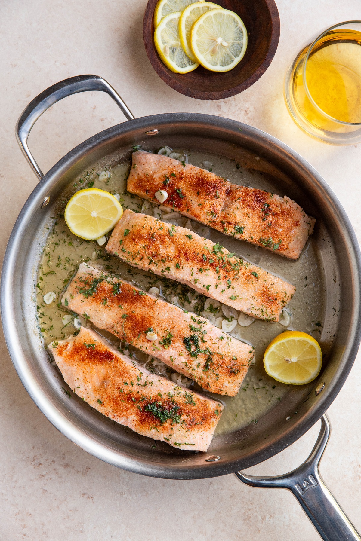 Poached salmon in a stainless steel skillet with a bowl of lemon slices and a glass of white wine to the side.