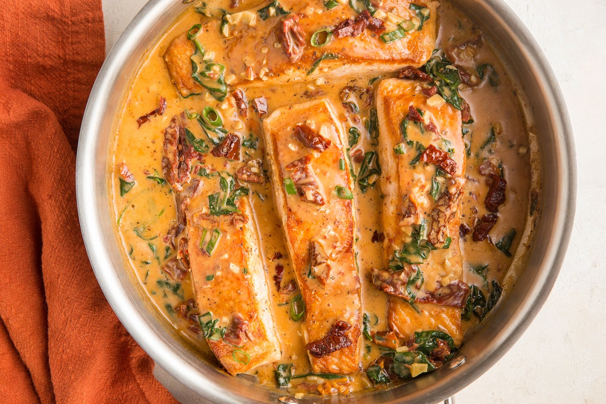 4 salmon filets covered in sun-dried tomato cream sauce in a skillet with a red napkin to the side.