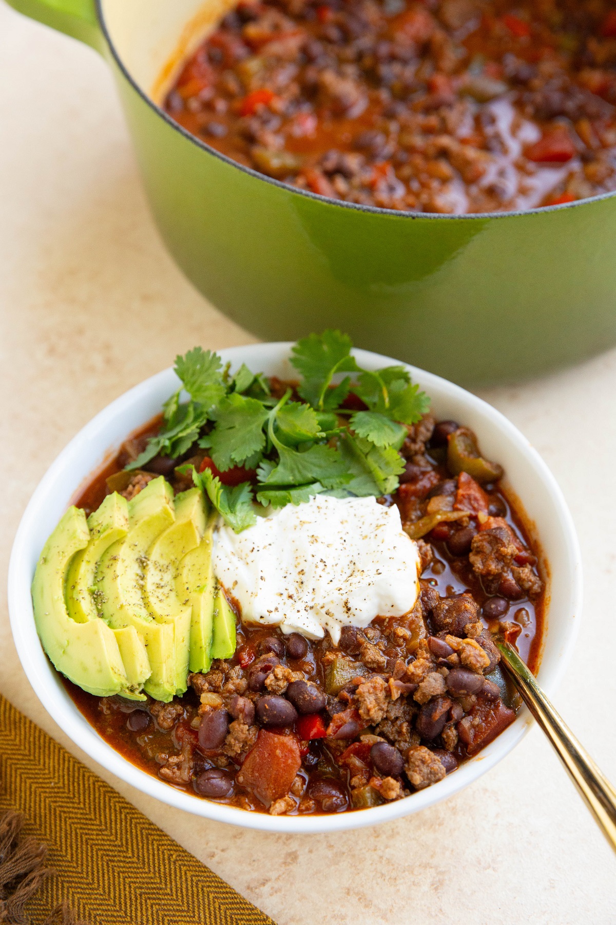 Big bowl of deer chili with sour cream, cilantro and sliced avocado on top with a pot of chili in the background.