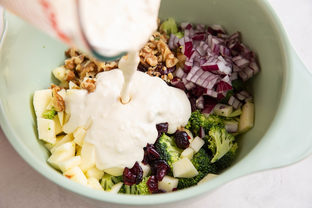 Pouring creamy dressing into the bowl with the broccoli salad ingredients.