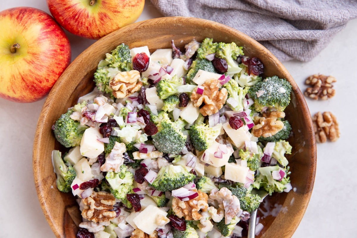 Broccoli salad in a wooden bowl with fresh apples and walnuts to the side.