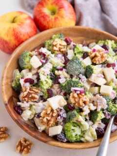 Wooden bowl of broccoli salad with a serving spoon, and fresh apples in the background.