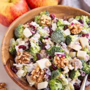 Wooden bowl of broccoli salad with a serving spoon, and fresh apples in the background.