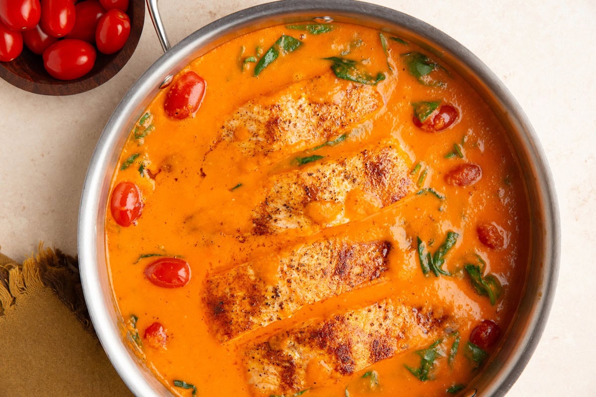 Stainless steel skillet with four salmon fillets cooked in roasted red pepper sauce.